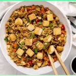 A picture of 15-Minute Meal Prep Cauliflower Fried Rice With Crispy Tofu in a bowl with a pair of chopsticks in it. A are of soy sauce is beside it.