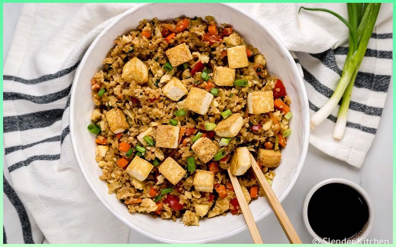 A picture of 15-Minute Meal Prep Cauliflower Fried Rice With Crispy Tofu in a bowl with a pair of chopsticks in it. A are of soy sauce is beside it.