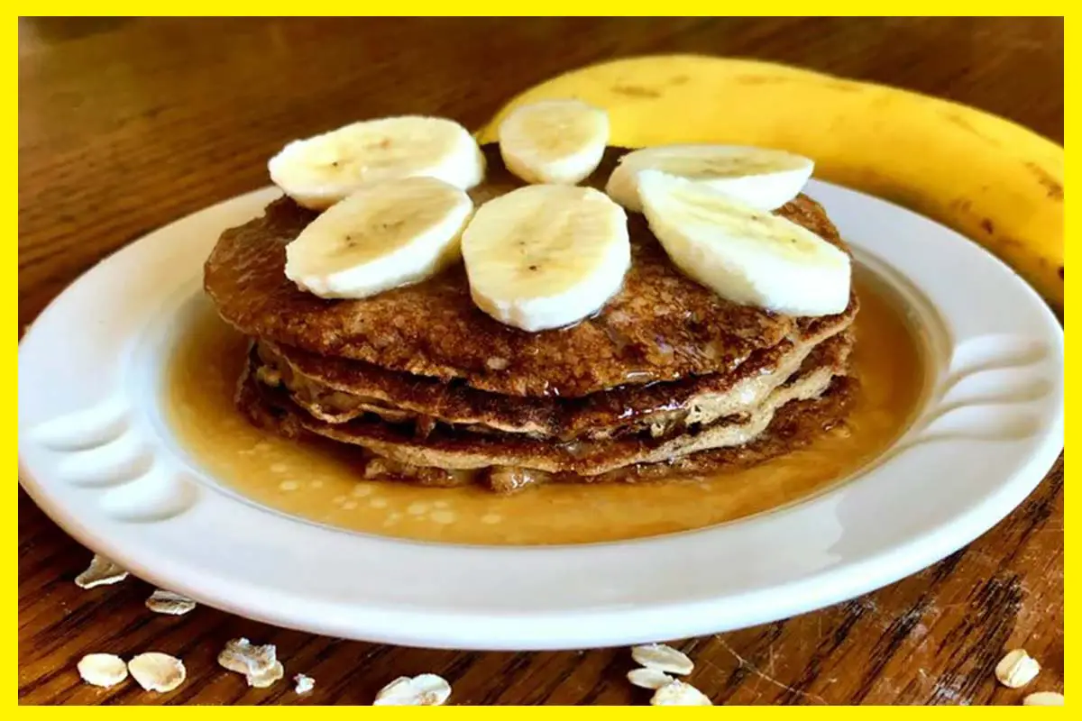 A picture of Almond Oat Banana Crepes with pieces of banana on it on white plate. A banana is by the side and ost are sprinkled on the brown table.